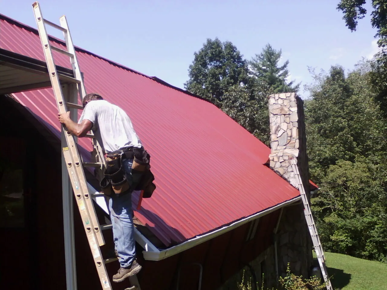 Roofer on a ladder beside a red metal roof and stone chimney — Soffit & Fascia Repair by Roofing Brothers in Ducktown